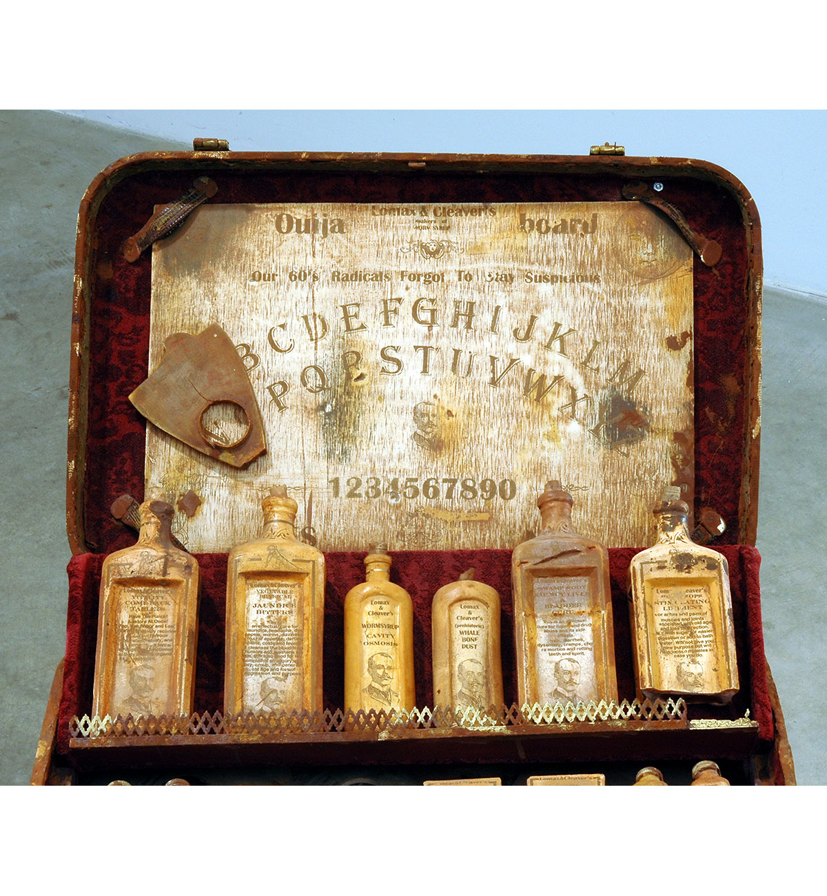 Dario Robleto
"Hippies And A Ouija Board (Everyone Needs To Cling To Something)", 2003-2004
Suitcase: cast and carved dehydrated bone calcium and bone dust from every bone in the body, microcrystalline cellulose, cold cast iron and brass, rust, antique syringe, crushed velvet, leather, thread, water extendable resin, typeset 
Bottles, medicines and Ouija board: cast and carved dehydrated bone calcium and bone dust from every bone in the body, typeset, homebrewed moonshine (potato derived alcohol), wine health tonics (water, sugar, fermented black cherries, yeast, gelatin, tartaric acid, pectinase, sulfur dioxide, oak flavoring, fortified with 100 year old: hemlock oil, Devil’s Claw, witch hazel bark, swamp root, powdered rhubarb, pleurisy root, belladonna root, white pine tar, coal tar, dandelion, sarsaparilla, mandrake, mullein, scullcap, cramp bark, elder, ginseng, horny goat weed, tansy, sugar of lead, mercury with chalk and tin-oxide; calcium, potassium, creatine, zinc, iron, nickel, copper, boron, vitamin k, crushed amino acids, home-cultured antibiotics, chromium, magnesium, colostrum, ironized yeast, ground pituitary gland, ground wisdom teeth, ground sea horse, shark cartilage, coral calcium, iodine and castor oil)
Records: various 1960’s 45 rpm records cast in prehistoric whale bone dust, typeset
23" x 19" x 42"