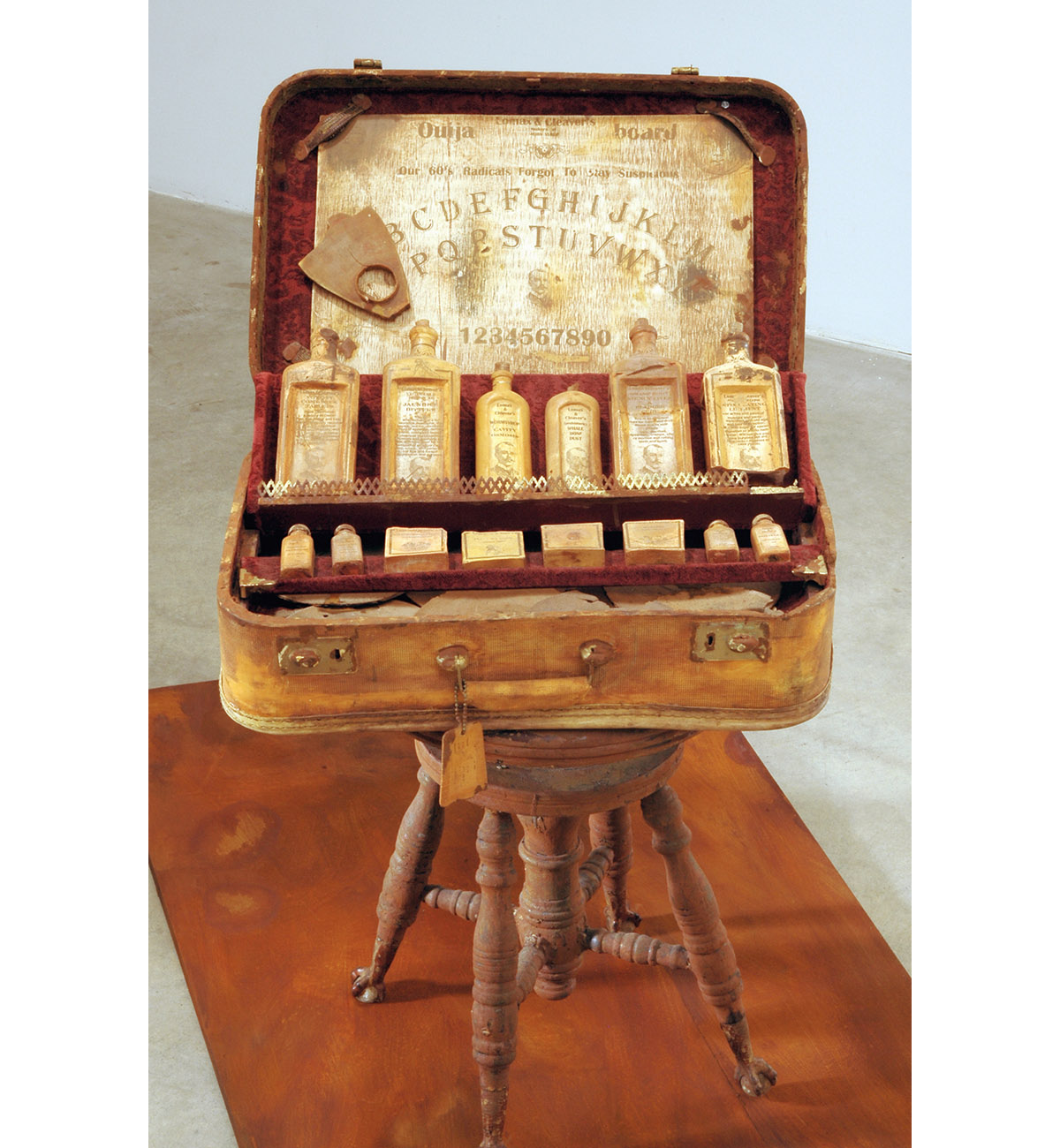 Dario Robleto
"Hippies And A Ouija Board (Everyone Needs To Cling To Something)", 2003-2004
Suitcase: cast and carved dehydrated bone calcium and bone dust from every bone in the body, microcrystalline cellulose, cold cast iron and brass, rust, antique syringe, crushed velvet, leather, thread, water extendable resin, typeset 
Bottles, medicines and Ouija board: cast and carved dehydrated bone calcium and bone dust from every bone in the body, typeset, homebrewed moonshine (potato derived alcohol), wine health tonics (water, sugar, fermented black cherries, yeast, gelatin, tartaric acid, pectinase, sulfur dioxide, oak flavoring, fortified with 100 year old: hemlock oil, Devil’s Claw, witch hazel bark, swamp root, powdered rhubarb, pleurisy root, belladonna root, white pine tar, coal tar, dandelion, sarsaparilla, mandrake, mullein, scullcap, cramp bark, elder, ginseng, horny goat weed, tansy, sugar of lead, mercury with chalk and tin-oxide; calcium, potassium, creatine, zinc, iron, nickel, copper, boron, vitamin k, crushed amino acids, home-cultured antibiotics, chromium, magnesium, colostrum, ironized yeast, ground pituitary gland, ground wisdom teeth, ground sea horse, shark cartilage, coral calcium, iodine and castor oil)
Records: various 1960’s 45 rpm records cast in prehistoric whale bone dust, typeset
23" x 19" x 42"