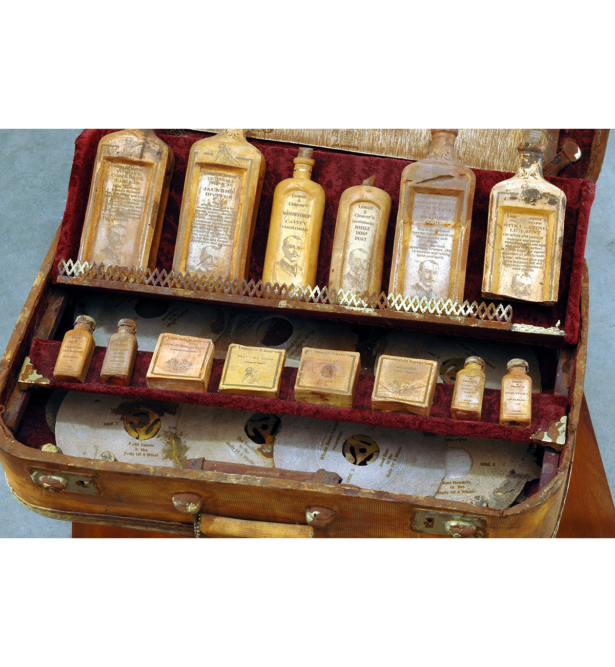 Dario Robleto
"Hippies And A Ouija Board (Everyone Needs To Cling To Something)", 2003-2004
Suitcase: cast and carved dehydrated bone calcium and bone dust from every bone in the body, microcrystalline cellulose, cold cast iron and brass, rust, antique syringe, crushed velvet, leather, thread, water extendable resin, typeset 
Bottles, medicines and Ouija board: cast and carved dehydrated bone calcium and bone dust from every bone in the body, typeset, homebrewed moonshine (potato derived alcohol), wine health tonics (water, sugar, fermented black cherries, yeast, gelatin, tartaric acid, pectinase, sulfur dioxide, oak flavoring, fortified with 100 year old: hemlock oil, Devil’s Claw, witch hazel bark, swamp root, powdered rhubarb, pleurisy root, belladonna root, white pine tar, coal tar, dandelion, sarsaparilla, mandrake, mullein, scullcap, cramp bark, elder, ginseng, horny goat weed, tansy, sugar of lead, mercury with chalk and tin-oxide; calcium, potassium, creatine, zinc, iron, nickel, copper, boron, vitamin k, crushed amino acids, home-cultured antibiotics, chromium, magnesium, colostrum, ironized yeast, ground pituitary gland, ground wisdom teeth, ground sea horse, shark cartilage, coral calcium, iodine and castor oil)
Records: various 1960’s 45 rpm records cast in prehistoric whale bone dust, typeset
23" x 19" x 42"